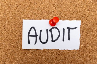 AUDIT 'nin iş ortamında mali inceleme ve değerlendirmeyi sembolize eden kırmızı raptiyeli bir mantar panoya tutturulduğunu belirten elle yazılmış bir not..