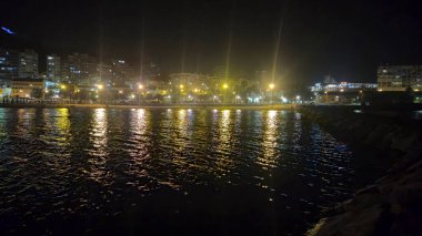Alicante İspanya - 17 Temmuz 2025: Aydınlatılmış binalar ve su üzerindeki yansımaları ile Alicante limanının gece manzarası. Şehir gece hayatı atmosferini vurgulayan sakin bir sahne.