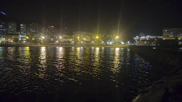 Alicante İspanya - 17 Temmuz 2025: Aydınlatılmış binalar ve su üzerindeki yansımaları ile Alicante limanının gece manzarası. Şehir gece hayatı atmosferini vurgulayan sakin bir sahne.