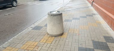 Yoğun bir şehir sokağının yanındaki ıslak kaldırımda beton bir çöp kutusu duruyor. Sahne, hareketli bir şehir yaşamında temiz ve kamusal alan kullanımını vurgulayan sakin bir anı yakalıyor..