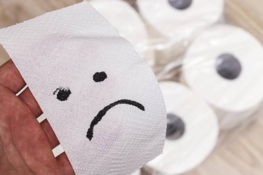 Bir el, arka planda tuvalet kağıdı ruloları ile çevriliyken hayal kırıklığını simgeleyen üzgün bir yüz çizilmiş bir kağıt havlu tutuyor..