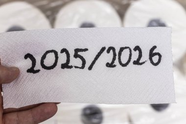 Üzerinde 2025 / 2026 sayıları yazılı bir kağıt parçasını tutan ve arka planda bulanık tuvalet kağıtları olan bir el, gelecek planları ve tuvalet kağıdı kullanımını temsil ediyor..