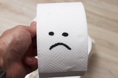 Üzücü bir yüz çizilmiş tuvalet kağıdı rulosunu tutan bir el. Bu görüntü, günlük nesnelerde mizah ve yaratıcılığı çağrıştırıyor. Sahne parlak ve basit..