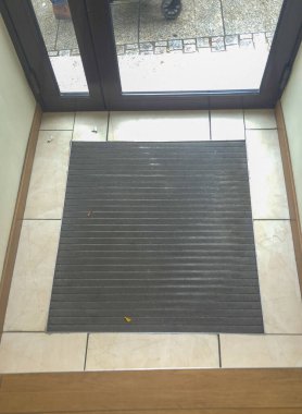 Kiremitli karşılama ziyaretçilerinin üzerine desenli lastik bir paspas yerleştirilmiş kapalı bir giriş. Doğal ışık, cam kapıdan süzülerek girişteki atmosferi arttırır..