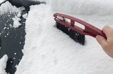 Arabanın çatısındaki karı temizlemek için kar fırçası kullanan bir el. Sahne, araba sürmeden önce kar birikimini ve araç bakımını vurgulayan kış havasını yakalıyor.