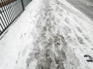 Soğuk bir kış gününde yürüyen buz ve insanların ayak izlerinin yer aldığı karlı kaldırım kentsel kış manzarasını ve havanın günlük hayata etkilerini vurguluyor..