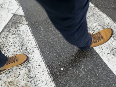 Yaya geçidinde yürüyen, kahverengi çizmeli ve pantolonlu bir adam. Sahne, kentteki hareketliliği ve günlük yaşamı görüntülüyor..