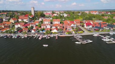 Masuria 'nın pitoresk bir kasabası olan Mikolajki' nin hava manzarasında, parlak bir gökyüzü altında, kayıklarla, büyüleyici evlerle ve manzaralı yeşilliklerle dolu canlı bir marina sergileniyor..