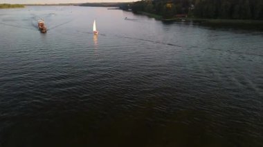 Mikolajki, Polonya, 9 Ağustos 2024: Daha büyük bir teknenin yanında süzülen yelkenli göl manzarası. Sakin sular, güneş ışığı parıldarken etrafındaki ağaçları yansıtır.