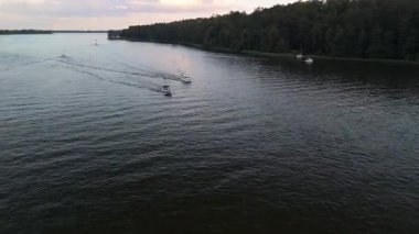 Mikolajki, Polonya, 9 Ağustos 2024: Tekneler, Masuria 'nın el değmemiş sularında pürüzsüz bir şekilde seyrederek, koyu renkli bir gökyüzü altında kıyı şeridi boyunca canlı yeşillikler sergiliyor.