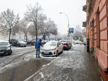 Rzeszow Polonya - 21 Kasım 2025: Kar yağışı, şehir ortamında kış havasını gösteren park halindeki bir arabadan kar temizleyen bir adamın şehir caddesini kaplaması.