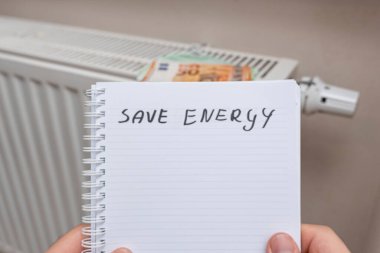 El yazısıyla yazılmış bir not, SAVAŞ ENERGY 'nin bir radyatörün önünde tutulduğunu ve sürdürülebilirlik için ev ısıtmasında enerji korumanın önemini vurguluyor..