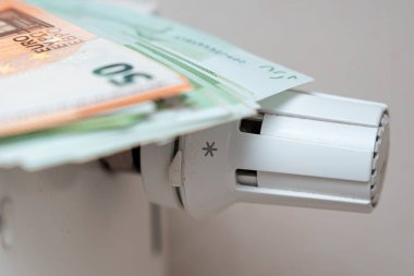 Konfor ve giderlerin dengesini vurgulayan ev ısıtma sistemleri içindeki finansal yönetim ve bütçeyi temsil eden beyaz bir radyatör vanasının üzerine yerleştirilmiş birkaç Euro banknotunun yakın plan görüntüsü.