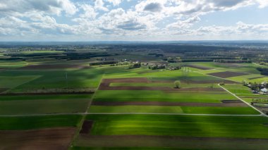 Parçalı bulutlu bir gökyüzünün altındaki yemyeşil tarlaların ve tarım arazilerinin havadan görünüşü kahverengi ve yeşil parçalarla dolu canlı bir kırsal manzarayı gösteriyor. Tarım temaları için ideal.