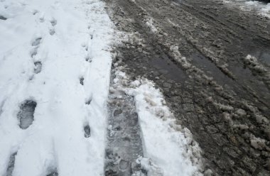 Karlı bir patika, kışın bahara geçişini gösteren çamurlu izlerle karışıyor. Ayak izleri birbirine zıt dokularda ilerleyip buzlu ama dünyevi bir sahneyi yakalıyor..
