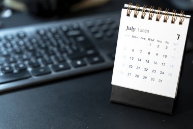Bir masaüstü takvimi Temmuz 2026 'yı gösteriyor ve bir bilgisayar klavyesinin yanına konmuş spiral bir bağlayıcı yerleştirilmiş. Takvim, zaman yönetiminin net bir görüntüsünü sunan günleri vurgular.
