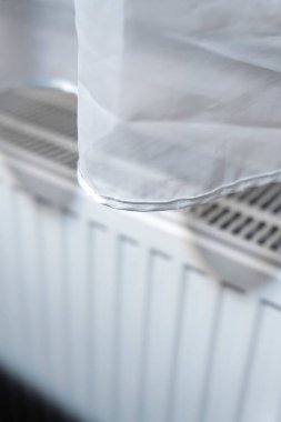 Sıcak bir odadaki radyatörün üzerine zarif bir şekilde örtülmüş beyaz bir perdenin yakın çekimi. Yumuşak kumaş, ısıtma ünitesinin zarif tasarımıyla çelişiyor ve evde sakin bir atmosfer yaratıyor..