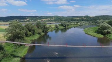 Hafif yüzey dalgalanmaları ve çevresindeki yeşilliklerle nehri geçen bir asma köprünün geniş hava görüntüsü güneşli koşullar altında dengeli bir kırsal alan oluşturuyor.