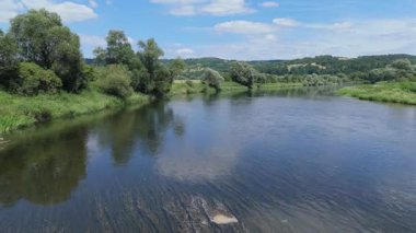 Durgun bir nehrin üzerinde, sakin bir su akıntısı ve bereketli bir yaz güneşi altında yemyeşil bankalarla asılı duran bir yaya köprüsünün uzak hava görüntüsü.