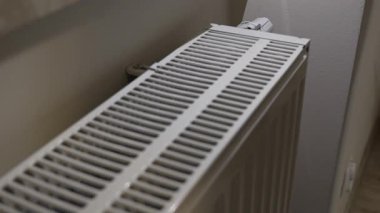 Bir ev kaloriferini yakından gören bir kişi, ısıyı kontrol etmek için ellerini ızgaranın üstüne koyar. Isıtıcının içerideki ısıtma sistemi kullanımı ve yerli enerji tüketimini vurguluyor.