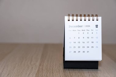 Aralık 2026 'da vurgulanmış tarihleri gösteren bir masa takviminin yakın çekimi. Takvim, ofis dekorasyonu için mükemmel işlevsellik ve estetiği harmanlayan ahşap bir yüzeye yerleştirilmiştir..