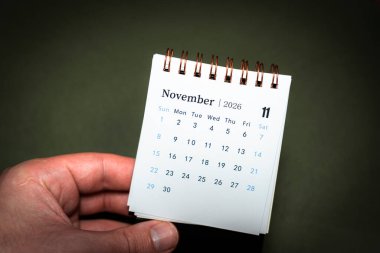 Kasım 2026 'yı gösteren spiral bir masa takvimi tutan bir el. Bu resim zaman yönetimi ve planlama kavramlarını etkili bir şekilde yakalar.