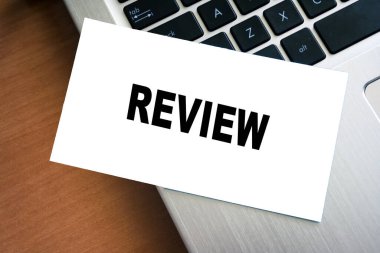 Dizüstü bilgisayarın klavyesine yerleştirilmiş beyaz bir kağıtta REViEW sözcüğünü gösteren el yazısıyla yazılmış bir not. Profesyonel ortamlarda geri bildirim ve değerlendirme ile ilgili kavramlar için ideal.