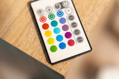 Renkli bir LED uzaktan kumanda, ışıklandırma efektlerini özelleştirmek için çeşitli renk düğmelerini gösteren ahşap bir yüzeye yerleştirilir. Ev dekorasyonu atmosfer ve teknoloji senaryoları için ideal..