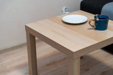 Mavi bir kupa ve beyaz bir tabak sergilenen ahşap bir masa oturma odasında minimalist bir ortam yaratıyor. Ev dekorasyonu temalı projeler veya yaşam tarzı içeriği için ideal.