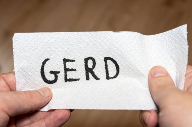 GERD sözcüğünü kağıt havlu üzerinde gösteren el yazısıyla yazılmış bir not. Genel bir sağlık durumunu simgeliyor. Bağlam, gastroözofageal reflüyle ilgili sağlık tartışmaları veya bilgilendirme materyalleri önermektedir..