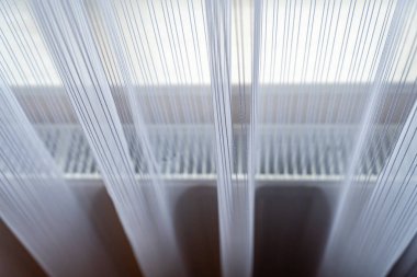 Beyaz bir radyatörün yanında yumuşak bir esintide sallanan narin perdelerin yakın çekim görüntüsü. Işığın etkileşimi odaya sakinleştirici bir etki yaratıyor..