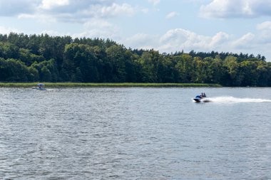 Mikolajki Polonya - 29 Eylül 2024: İki tekne, bulutlu bir gökyüzü altındaki yemyeşil ağaçlarla çevrili sakin bir gölde geziniyor ve boş zamanları ve doğa aktivitelerini vurguluyor.