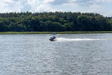 Mikolajki Polonya - 29 Eylül 2024: Su sporları ve eğlence faaliyetlerini sembolize eden berrak bir gökyüzünün altında yemyeşil arka planda sakin bir gölde hızla ilerleyen jet ski.