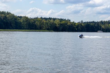 Mikolajki Polonya - 29 Eylül 2024: Bireylerin yemyeşil ormanlar ve açık mavi gökyüzü ile çevrili sakin bir göl üzerinde jet ski sürüşünün keyfini çıkardıkları heyecan verici bir an.