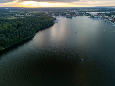 Aerial view of a serene lake surrounded by lush forests and a small town at sunset. The water reflects warm colors as boats navigate the calm surface.