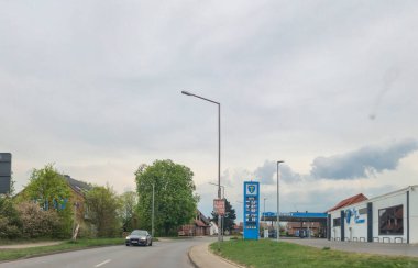 Almanya - 15 Nisan 2025: Almanya 'da küçük bir kasabanın atmosferini aydınlatan ağaçlar ve binalarla çevrili dolambaçlı bir yolda bir arabanın yer aldığı şehir sahnesi.