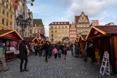 Wroclaw Polonya - 3 Ocak 2026: Ürünlerin arka planında tarihi binalar ve caddeyi aydınlatan şenlikli dekorasyonlar keşfeden hareketli alışveriş merkezleriyle dolu kış piyasa sahnesi.
