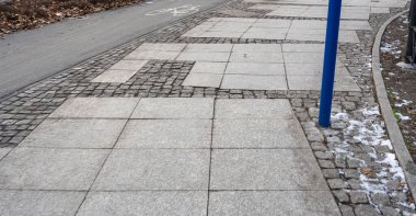 Yumuşak beton ve kaldırım taşlarının karışımından oluşan bir kaldırımın yakın çekimi. Sahne, çağdaş kamusal alan gösteren mavi bir direkle kentsel tasarımı yakalıyor..