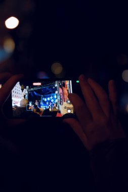 Sahnede canlı bir performans yakalayan bir kişi, renkli ışıklarla aydınlatılmış canlı bir şehir ortamında akıllı bir telefonla festival atmosferi sergiliyor..