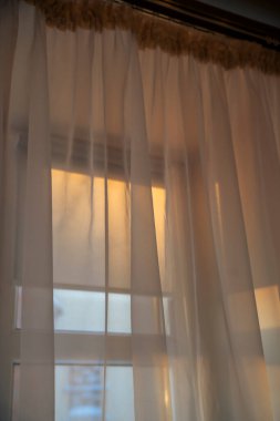 Perdeler rüzgarda hafifçe sallanarak güneşli bir pencere sahnesini açığa çıkarıyor. Kumaştan gelen sıcak ışık geçişleri kapalı bir atmosfer yaratıyor..