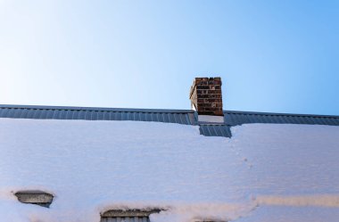 Tuğla bacalı karla kaplı bir çatı açık mavi bir gökyüzüne karşı duruyor. Kar birikimi, sakin bir kırsal ortamı ön plana çıkaran kış koşullarını sergiliyor.
