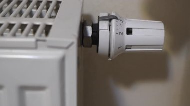 İnsan elinin beyaz bir radyatör termostat topuzunu içeride döndürdüğü yakın çekim görüntüsü. Parmaklar valfi yavaşça çevirirken kamera sabit kalır, sakin bir ev ısıtma sahnesinde elle ısı ayarlamasını gösterir.