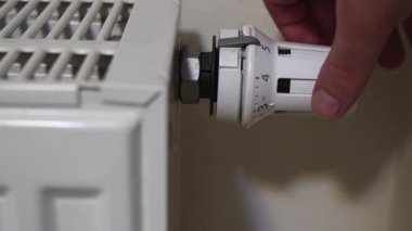 Bir odanın içinde radyatör termostat vanasını çeviren bir elin makro yakın çekimi. Kamera, parmaklar ısıtma seviyesini ayarlarken sabit kalıyor. Bu da yerleşim yerindeki basit bir elle kontrolü gösteriyor..