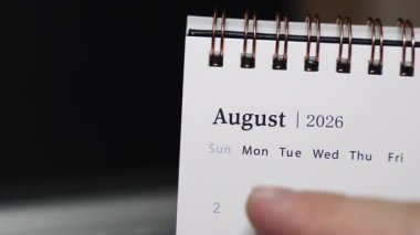Laptop klavyesindeki bir masa takviminin insan parmağı olarak yakın plan görüntüsü Ağustos 2026 'daki belirli bir tarihe işaret ediyor. Statik kamera modern bir çalışma alanında kesin tarih göstergesini ve aylık yapılandırılmış planlamayı vurguluyor.