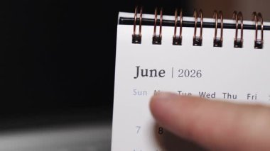 Bir dizüstü bilgisayar klavyesine insan parmağı olarak yerleştirilen bir masa takviminin yakın çekimi Haziran 2026 'daki belirli bir tarihi gösteriyor. Statik kamera modern bir çalışma alanında kesin tarih seçimi ve aylık planlama kavramını vurguluyor.