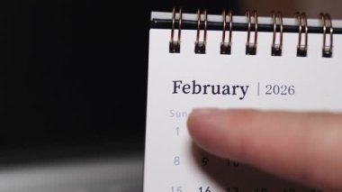 Bir dizüstü bilgisayarın klavyesindeki bir masa takviminin insan parmağı olarak yakın plan görüntüsü Şubat 2026 'daki belirli bir tarihi gösteriyor. Statik kamera, modern bir çalışma alanında kesin tarih seçimi ve temiz aylık planlama kavramını vurgular.