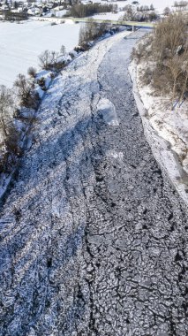 Kışın gözlemlenen karla kaplı donmuş bir nehrin havadan görünüşü. Buz oluşumları ve karla kaplı bankalar kırsal bir ortamda soğuk, sakin bir manzara yaratıyor..