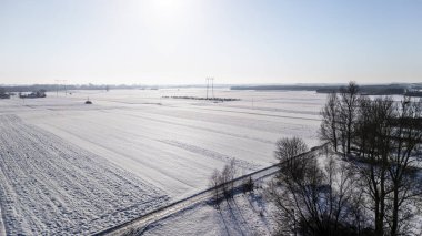 Buzla kaplı tarlaları ve açık mavi gökyüzüyle karla kaplı engin bir arazinin havadan görünüşü. Sahne, kırsal kesimde kışın dinginliğini vurguluyor ve uzaktaki elektrik hatlarını gösteriyor..