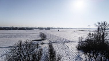 Güneşin aydınlattığı açık mavi gökyüzünün altında geniş karla kaplı tarlaların ve uzak ağaçların bulunduğu sakin bir kış manzarası. Doğa sükuneti ve mevsimlerle ilgili projeler için ideal.