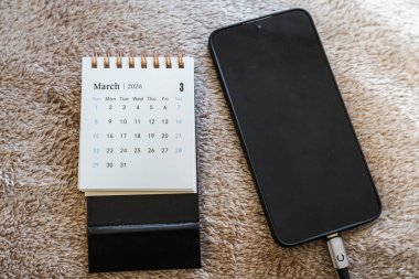 Mart 2026 'yı gösteren bir takvim lüks bir yüzeyde modern bir akıllı telefonun yanında duruyor. Telefon teknoloji ve bağlantıyı temsil ederken takvim önemli tarihleri gösterir.